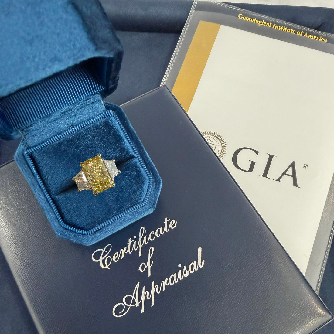 Completed custom three-stone emerald cut engagement ring in box with certificate of appraisal.