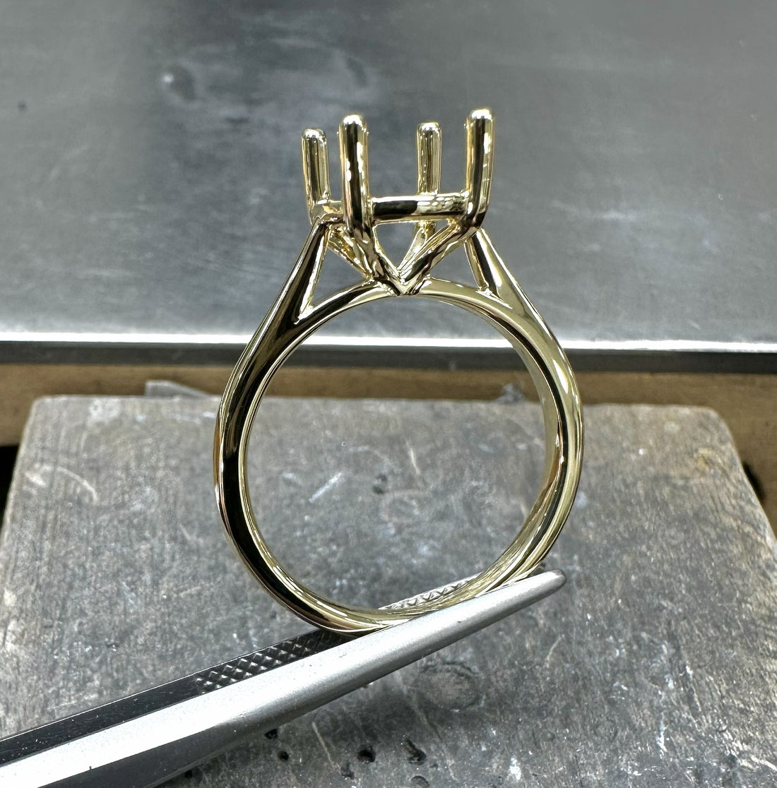A gold four-prong ring setting held upright on a workbench.