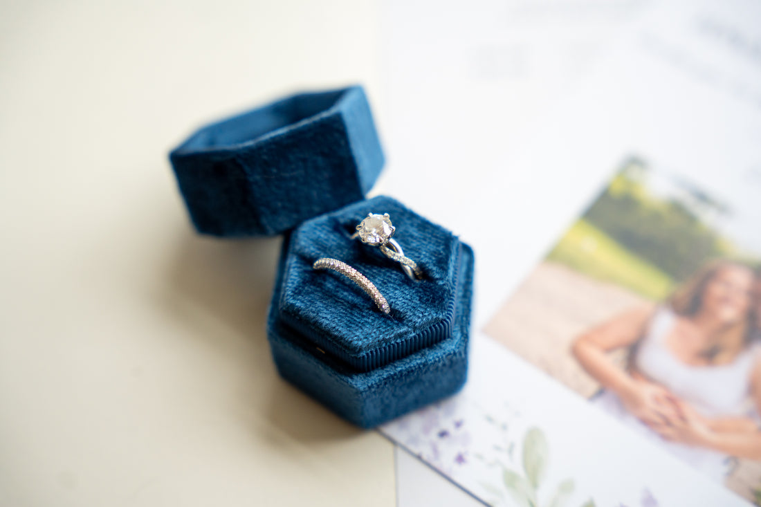 An open dark blue velvet ring box, holding an engagement ring with a round diamond with a twist shank and a three-sided wedding band. A blurred engagement photo is visible in the background.