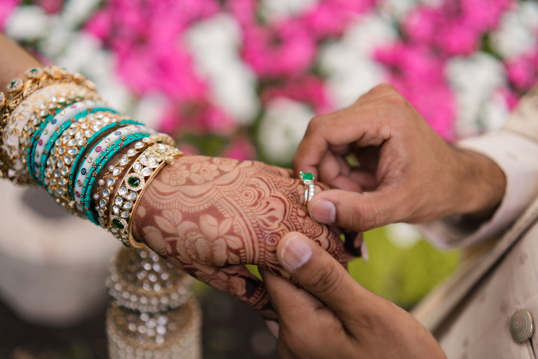 A close-up of a hand with intricate henna designs and stacked turquoise, white, and gold bangles, receiving a wedding band.