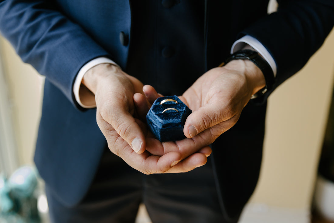 A close-up of a groom in a navy blue suit holding a small dark blue velvet ring box with two gold wedding rings inside.