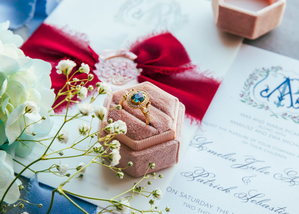 A yellow gold ring, featuring an oval Montana Sapphire displayed on a pink velvet ring box, surrounded by white flowers, a red ribbon, and wedding stationery.