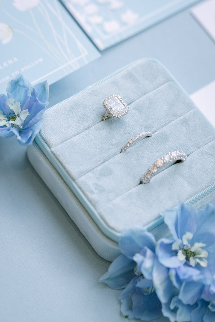 Three white gold rings, including a clawless halo engagement ring, and wedding bands, displayed on a light blue velvet jewelry box, surrounded by blue flowers.