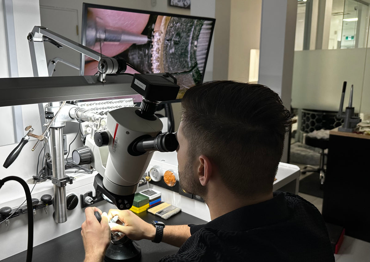 Patrick working on micropave through microscope at a workbench. A large monitor above displays a close-up view of the work