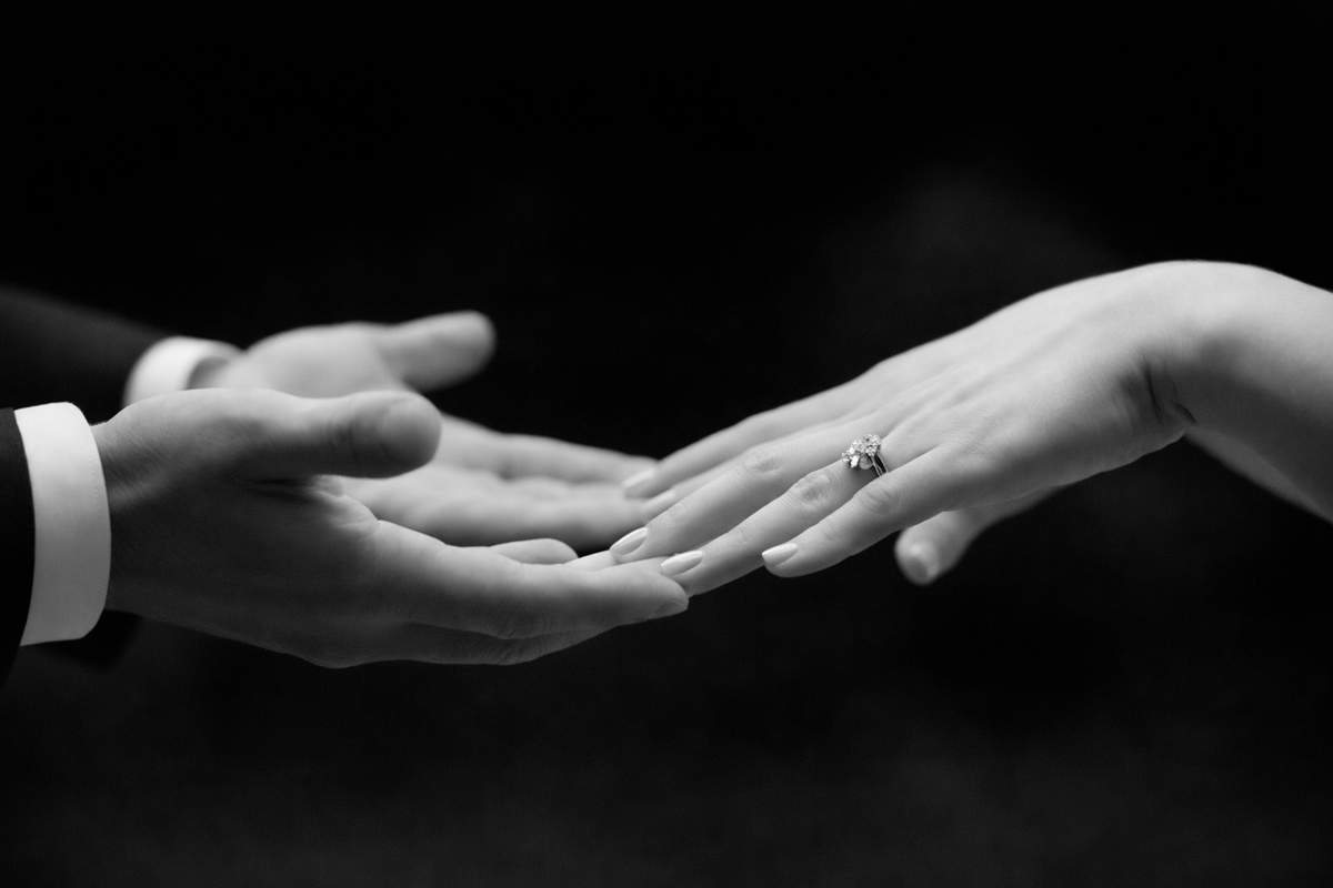 Black and white close-up of two hands reaching toward each other at a wedding, with an engagement ring worn.