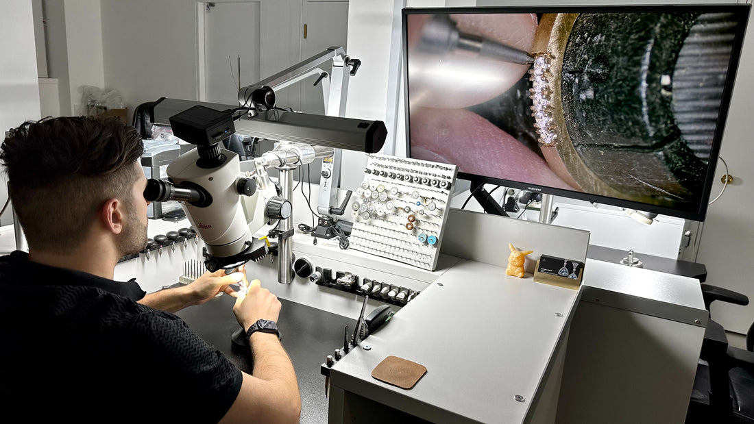 A jeweler working at a microscope and computer monitor in a workshop, with a screen showing a ring being set.