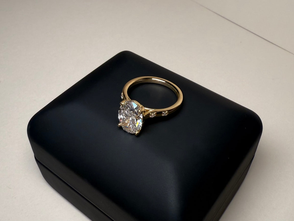 A yellow gold ring with a large oval diamond and small side stones accents, displayed on a black jewelry box.