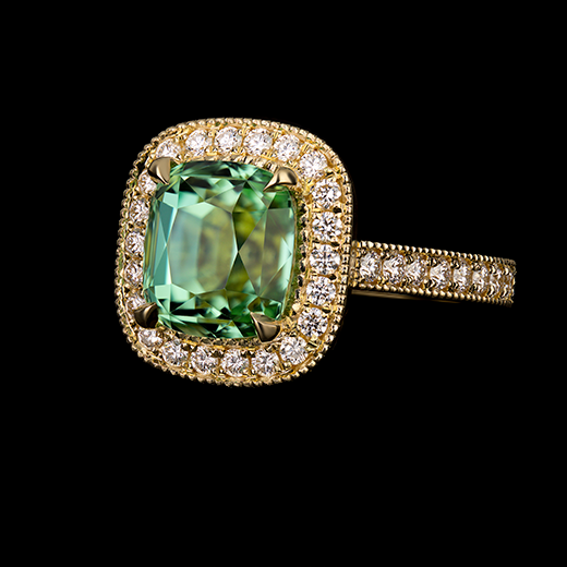 3.35 CT cushion cut Tourmaline center stone set in four crisp talon claws. Surrounded by round brilliant cut diamonds in the halo with a hand-applied mill grain edge. Accented by round diamonds cascading down the shank with a hand-applied mill grain edge. The stunning ring is made in 18k yellow gold.