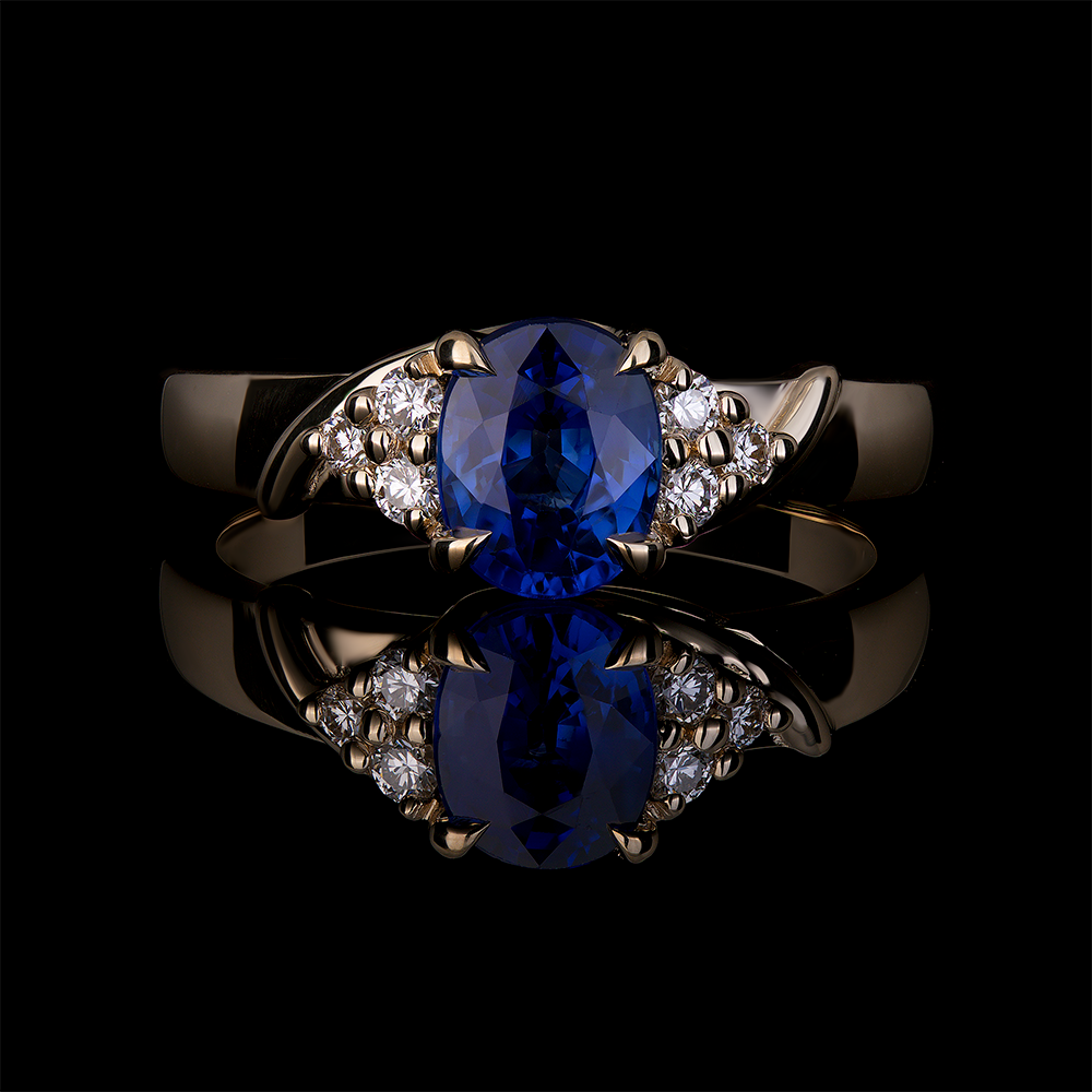 1.22 CT Ceylon blue oval sapphire center stone set in 14k yellow gold mount with 4 talon claws. Accented with 3 round diamonds on each shoulder set with round prongs.